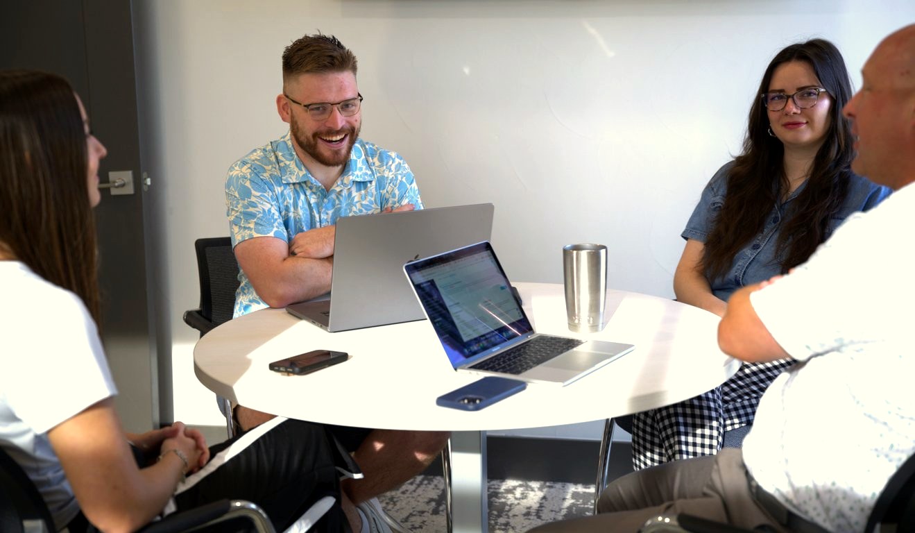 Four people talking around a round table with laptops in a bright meeting room